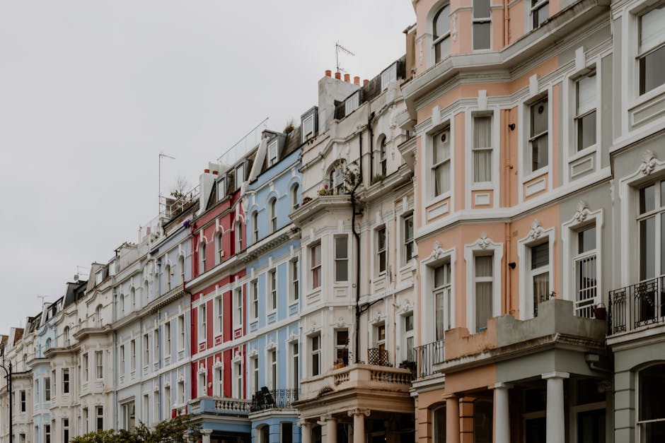 A row of Victorian terraced houses with ornate architectural details and large sash windows, painted in pastel shades of cream, pink, blue, and white. The buildings are four to five stories high, with some featuring small balconies with decorative wrought iron railings. The rooftops have chimneys and some have rooftop antennas or small rooftop gardens. The sky is overcast, creating diffused lighting that highlights the textures of the facades, including decorative moldings and window frames. The buildings are closely packed along a street with no visible vehicles or pedestrians. The overall scene depicts a typical identified residential area, illustrating the type of urban environment where professional waste management and private rubbish removal services may operate, such as those offered by House Clearance Kentish Town, to manage waste generated in city living spaces.