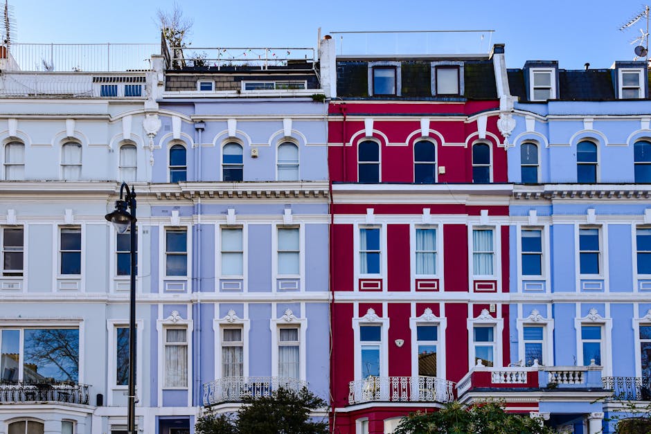 A row of Victorian terraced houses with ornate architectural details and large sash windows, painted in pastel shades of cream, pink, blue, and white. The buildings are four to five stories high, with some featuring small balconies with decorative wrought iron railings. The rooftops have chimneys and some have rooftop antennas or small rooftop gardens. The sky is overcast, creating diffused lighting that highlights the textures of the facades, including decorative moldings and window frames. The buildings are closely packed along a street with no visible vehicles or pedestrians. The overall scene depicts a typical identified residential area, illustrating the type of urban environment where professional waste management and private rubbish removal services may operate, such as those offered by House Clearance Kentish Town, to manage waste generated in city living spaces.