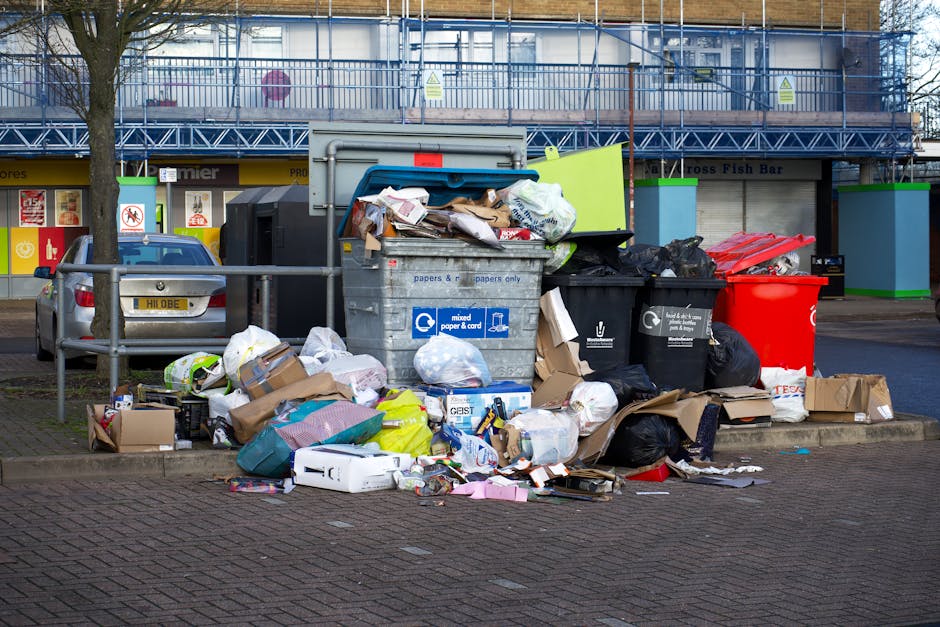 A large pile of mixed household waste and rubbish is accumulated outside a commercial building in a public parking area, including numerous black, black and white garbage bags, flattened cardboard boxes, crumpled paper, plastic packaging, and some unboxed items. The waste is situated next to a metal railing, with some items spilling onto the paved ground. Behind the waste, there are tall, multi-coloured structures, and further back, a building with a blue scaffolding or safety framework, indicating ongoing construction or maintenance work. A grey car is parked to the left of the rubbish, partially obscured by the railing. The scene appears to depict an instance of unmanaged or overflowing waste, possibly illustrating the need for independent waste collection services like those provided by House Clearance Kentish Town, especially for private or on-site clearance instead of council rubbish disposal. The environment is typical of an urban commercial or retail area, with storefronts and signage visible in the background, under an overcast sky with diffuse lighting.