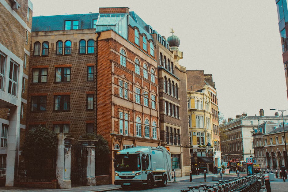A street scene featuring a mix of older and modern multi-storey buildings, with a prominent red brick structure in the center topped with a green dome and a small gilt cross. To the left, part of a white modern building with large glass windows is visible, while on the right, the facade of a yellow and cream Victorian-style building with detailed window frames can be seen. The street is lined with bollards and a row of bike racks in the foreground. A blue and white waste collection truck from House Clearance Kentish Town is parked on the street, partially blocking the view of the buildings behind it. The pavement appears clean, and the environment is well-lit, suggesting daytime, with overcast weather casting a soft, diffused light over the scene. This urban setting demonstrates an example of private waste collection activity amidst historic and contemporary architecture in the area, supporting alternative rubbish removal services that handle refuse directly on site.