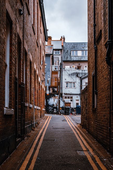 A narrow, cluttered back alleyway with uneven, muddy ground and scattered debris. On the right side, there are large white and beige plastic storage bins and a cardboard box, some of which appear to be used for waste storage or collection. Towards the foreground, a red plastic bucket is visible, and further down, multiple green wheelie bins are lined up against the left brick wall, some partially obscured by other objects. The alley is enclosed by weathered walls, one brick and one corrugated metal, both covered in graffiti with various tags and spray-painted designs. Electrical wires are strung overhead, connecting buildings on both sides of the alley, which also features wooden staircases leading to upper floors of adjacent buildings. The scene is lit by natural daylight, creating a slightly moody, urban atmosphere typical of environment where private rubbish collection or on-site waste disposal may occur, as handled by services such as House Clearance Kentish Town.