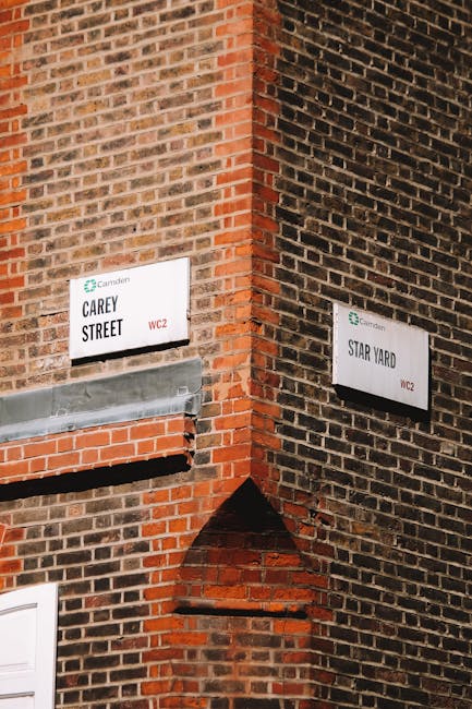 Two white street signs mounted on the corner of a historic brick building, one reading 'Carey Street' and the other 'Star Yard,' both indicating parts of Camden in Kentish Town. The Carey Street sign is positioned on the left side on a wall with a rough, weathered brick surface featuring various shades of red and brown, with a prominent gray metallic pipe or vent beneath it. The Star Yard sign is on the right side, attached to the darker brick wall, slightly higher, with a similar weathered texture. The building has a small, arched brick detail connecting the two facades, with the scene captured in natural daylight. The overall setting suggests an urban environment potentially involved with waste management or private property access, supported by the context of rubbish removal services. The image emphasizes traditional street signage in a gritty, historic setting, subtly reflecting an environment where alternative waste collection methods might be considered.