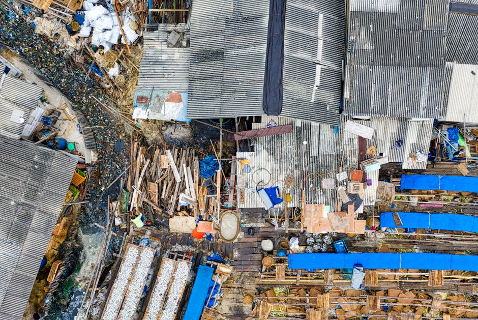 An aerial view of a narrow, cluttered outdoor area filled with various discarded materials, including wooden planks, plastic containers, metal sheets, and scattered debris. The scene features an arrangement of weathered, corrugated metal roofing panels in shades of grey and silver, some partially overlapping or leaning against structures. Wooden pallets and broken furniture are visible among the rubbish, with blue tarpaulins covering sections of the clutter. The environment appears to be a back yard or alley with limited space, surrounded by makeshift structures and sheds. Additionally, there are small piles of mixed waste, such as bottles and packaging, contributing to the overall disorder. The scene is uniformly lit, emphasizing the textures of the different materials, and highlights the challenge of managing waste in tight, congested urban settings. House Clearance Kentish Town occasionally handles such situations by offering private site clearance and rubbish removal services to clear clutter like this from narrow streets or small outdoor spaces, facilitating alternative waste handling outside local authority collections.
