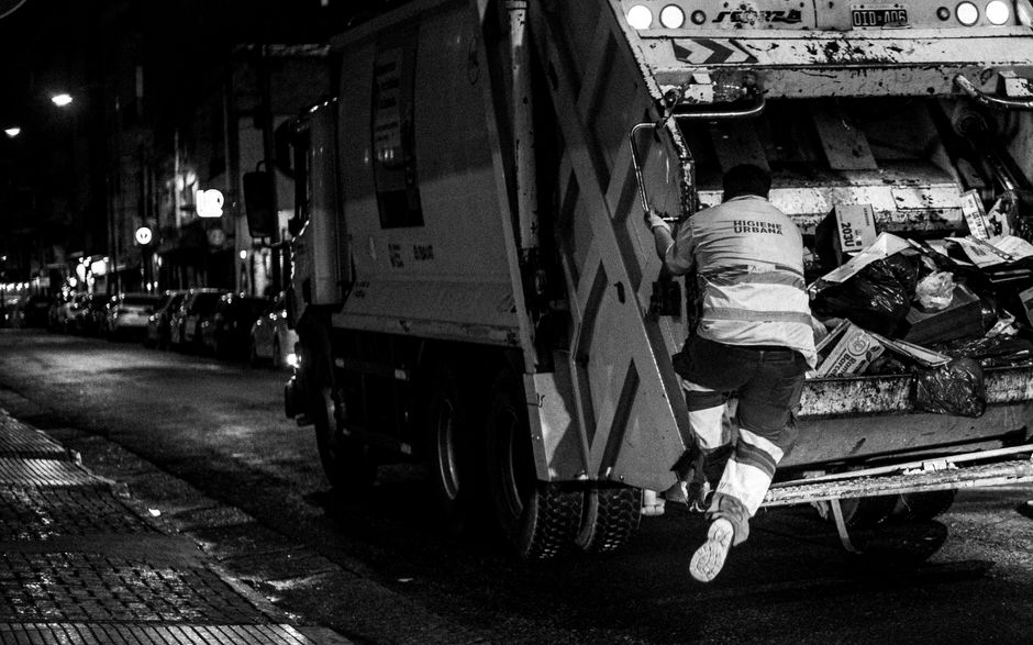 A worker wearing a high-visibility jacket with reflective strips is captured mid-action, climbing into the back of a large, industrial waste collection vehicle parked on a city street at night. The vehicle has a metallic, somewhat weathered exterior with visible dirt and grime, and its rear loading area is open, revealing a heap of mixed waste materials including black plastic bags, cardboard boxes, and various debris. The worker's trousers are dark with reflective bands, and their footwear is sturdy and white. The street surface appears wet, with reflections of nearby streetlights and parked cars lining the curb on the left side of the image. In the background, a row of cars is parked along the street, with some illuminated shop signs and streetlights creating a softly lit urban nighttime scene. This scene illustrates the process of private waste collection, possibly representing an alternative to council rubbish pickups, and emphasizes the role of professional rubbish removal services like House Clearance Kentish Town in managing urban waste efficiently and safely.