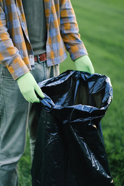 A person standing outdoors on a grassy area, wearing a checkered shirt with shades of yellow, blue, and gray over a green t-shirt, and gray pants secured with a brown belt, is holding open a large black heavy-duty rubbish bag lined with a shiny silver interior. The individual is wearing bright green disposable gloves, indicating they are engaged in waste collection or disposal. The background features well-maintained green grass, likely part of a garden or park setting. The scene suggests an activity related to rubbish removal or waste management, with the person preparing to discard garden debris, household waste, or other refuse. The focus is on the hand positioning and the bag, aligning with a professional waste collection service that handles on-site clearance. The setting appears clean and organized, emphasizing the importance of proper rubbish handling and disposal practices typically associated with independent or private waste management services. The natural daylight provides clear visibility of textures—such as the crinkled fabric of the bag and the smooth surface of the gloves—highlighting the practicality of the equipment used. This image visually supports topics related to rubbish removal, waste disposal regulations, and alternative waste handling processes outside of local authority collection, as seen in professional household clearance scenarios.