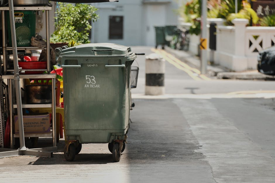A large green plastic wheeled rubbish bin with a slightly textured surface and a fitted lid, positioned on a paved sidewalk near the edge of a building with storage shelves containing various household items. The bin has white marking indicating the number '53' and the words 'JUN BESAR.' It is situated close to a curb and street, with the urban environment visible in the background, including a road with painted yellow lines, white concrete barriers, and a parked vehicle. The scene is brightly lit, suggesting daylight, and the surroundings include a residential area with fencing and greenery. This setup may relate to private waste collection or on-site clearance services provided by House Clearance Kentish Town, reflecting independent rubbish management rather than local authority collection.