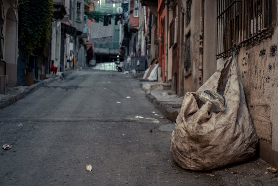 A collection of black garbage bags, cardboard boxes, and discarded packaging are piled against a weathered, off-white exterior wall below a red metal door covered with graffiti and stickers. The door features a small sign that reads 'No Money, Keep Clear' and another indicating 'Keep Clear,' suggesting restricted access. The rubbish is haphazardly arranged, with some bags leaning on the wall and others stacked nearby, indicating a private or independent rubbish collection outside a building. The area appears cluttered, with various materials including cardboard and plastic waste, typical of on-site rubbish clearance or waste disposal activities not managed by local authorities. The environment is illuminated by natural daylight, highlighting the textures of the garbage bags and the rough wall surface, with a narrow space in front of the door used for temporary waste accumulation, linking to waste management practices such as private waste removal services often used for house clearances or large waste loads in urban settings.