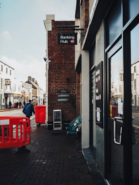 The image depicts a section of a street corner with a brick building featuring a black sign that reads 'Banking Hub,' positioned above a bank of large, dark glass windows with reflective surfaces. To the left, there is a red plastic barrier placed on the pavement, accompanied by a red waste bin. Near the building's wall, a small blackboard or notice board is positioned, possibly displaying information. In the background, pedestrians are walking along the street, with commercial buildings lining the opposite side, including shops with storefront windows and signs, all under a cloudy sky. The environment appears busy and urban, with typical street furniture and signage. The scene provides context for urban waste management and the potential role of private rubbish collection services, such as those offered by companies like House Clearance Kentish Town, in maintaining cleanliness in commercial and public areas.