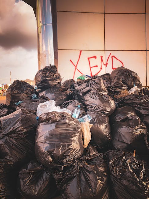 A large pile of black garbage bags, some with visible plastic bottles and waste materials, is stacked against a tiled wall in an outdoor urban setting. The bags are irregularly shaped, with some tightly knotted at the top and others loosely gathered, and they vary slightly in size. The wall behind features beige or light brown tiles with a subtle reflective finish, and red spray-painted graffiti spelling 'XEND' is visible on the tiling above the waste. The surrounding environment includes a partly cloudy sky with hints of sunlight and distant cityscape elements, indicating this waste collection scene is located outside a building, possibly associated with private waste handling or a rubbish clearance service. The overall scene reflects a typical instance of on-site rubbish removal, reminiscent of independent collection efforts or non-local authority disposal, and is managed by companies such as House Clearance Kentish Town that undertake rubbish clearance for various clients.