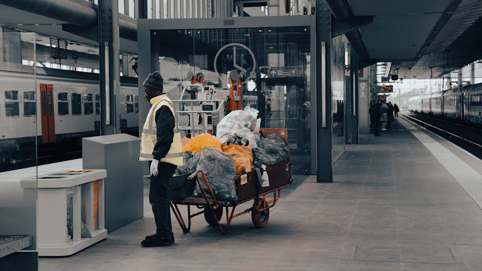 A man dressed in dark clothing, a black beanie, and a high-visibility vest stands next to a large metal trolley loaded with multiple black, grey, and orange plastic bags filled with waste at a modern train station platform. The trolley is positioned on the tiled surface adjacent to a white waste bin. Behind him, there are glass walls, metal structural elements, and a high-speed train on the right side of the image. In the background, other commuters are seen walking or waiting near the platform, and a train schedule display can be faintly seen. The scene is lit by natural daylight, lending a neutral tone to the setting, with the focus on the man and the waste bags, suggesting a scenario of waste collection or rubbish removal, perhaps in cooperation with private waste handling services like House Clearance Kentish Town. The station's contemporary architecture, with its clean, metallic, and glass features, provides a professional environment suitable for efficient rubbish collection activities happening away from typical residential or commercial areas.
