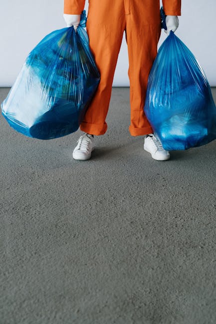 A person standing outdoors on a paved surface is holding two large blue plastic rubbish bags, one in each hand. The individual is dressed in orange trousers and white sneakers, with only the lower torso and legs visible. The bags are filled with waste and appear to be tied at the top, showing some compressed contents inside. The background is plain with a neutral-colored ground and a light gray wall, suggesting a driveway, parking area, or exterior space suitable for waste collection or disposal. The scene emphasizes the process of waste removal, reflecting a private or independent approach to rubbish collection, typical of services like those offered by House Clearance Kentish Town, which cater to on-site clearance needs and alternative waste handling methods. The focus remains on the physical act of carrying waste, supporting visual context related to rubbish removal and waste management activities.